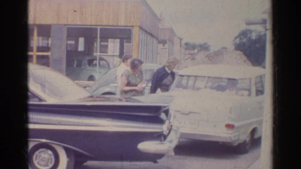 A woman bends slightly near the parked cars as another stands beside her at the 1961 German service-station scene.