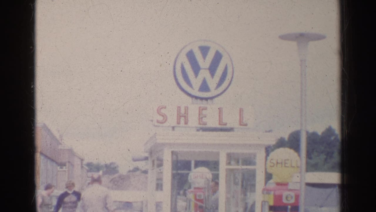 Shell service-station view in Germany with a Volkswagen logo sign above the building and people near cars at the lower edge.
