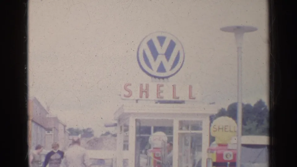 Shell service-station view in Germany with a Volkswagen logo sign above the building and people near cars at the lower edge.