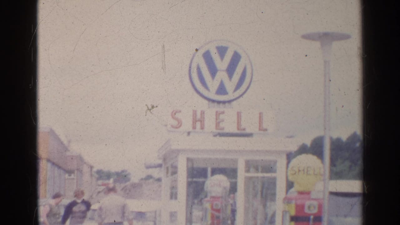 Shell station building with Volkswagen sign, pumps, cars, and adults on the forecourt.