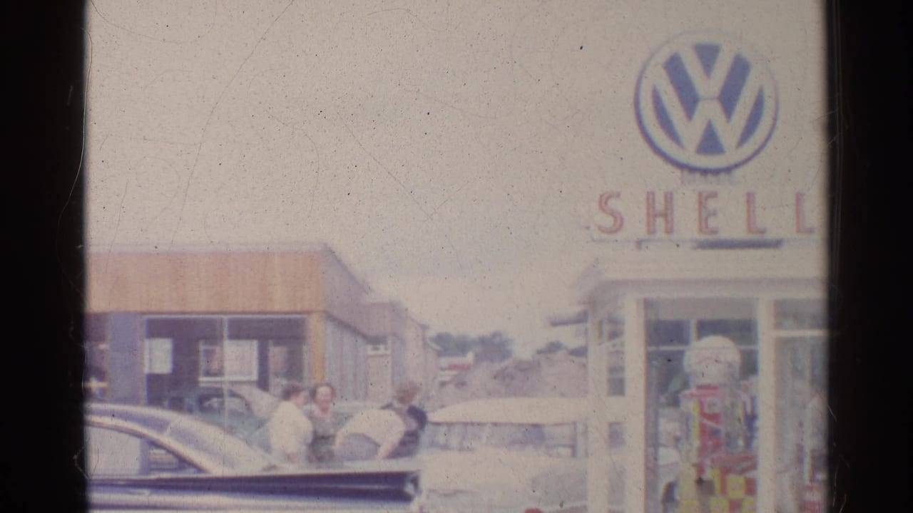 1961 Germany home-movie frame with adults near parked sedans, a Shell pump, and a partially framed Volkswagen sign on the station building.