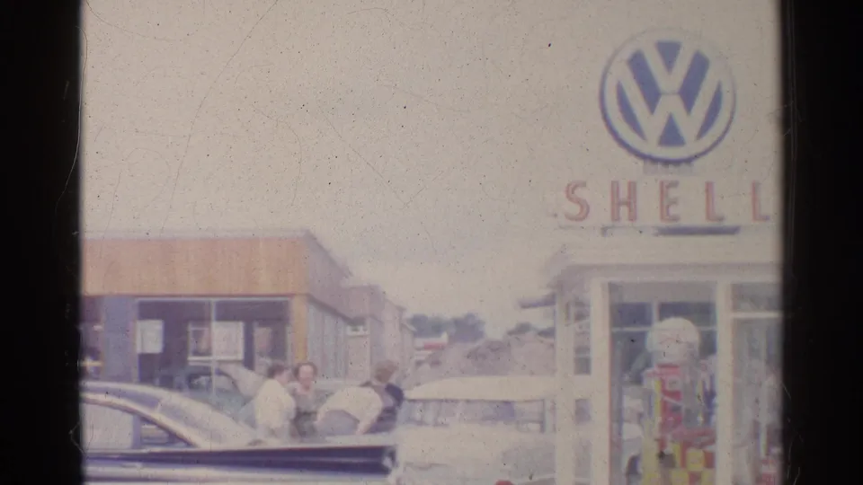 1961 Germany home-movie frame with adults near parked sedans, a Shell pump, and a partially framed Volkswagen sign on the station building.