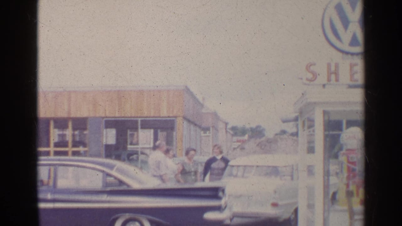 1961 Germany frame showing a dark sedan in the foreground, adults by a light car, a Shell pump, and cropped Volkswagen and Shell signage.