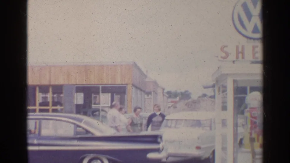 1961 Germany frame showing a dark sedan in the foreground, adults by a light car, a Shell pump, and cropped Volkswagen and Shell signage.