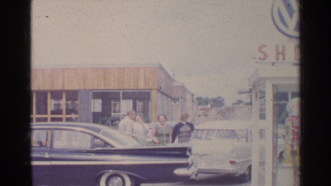 Adults stand between two parked cars outside a small gas station building.