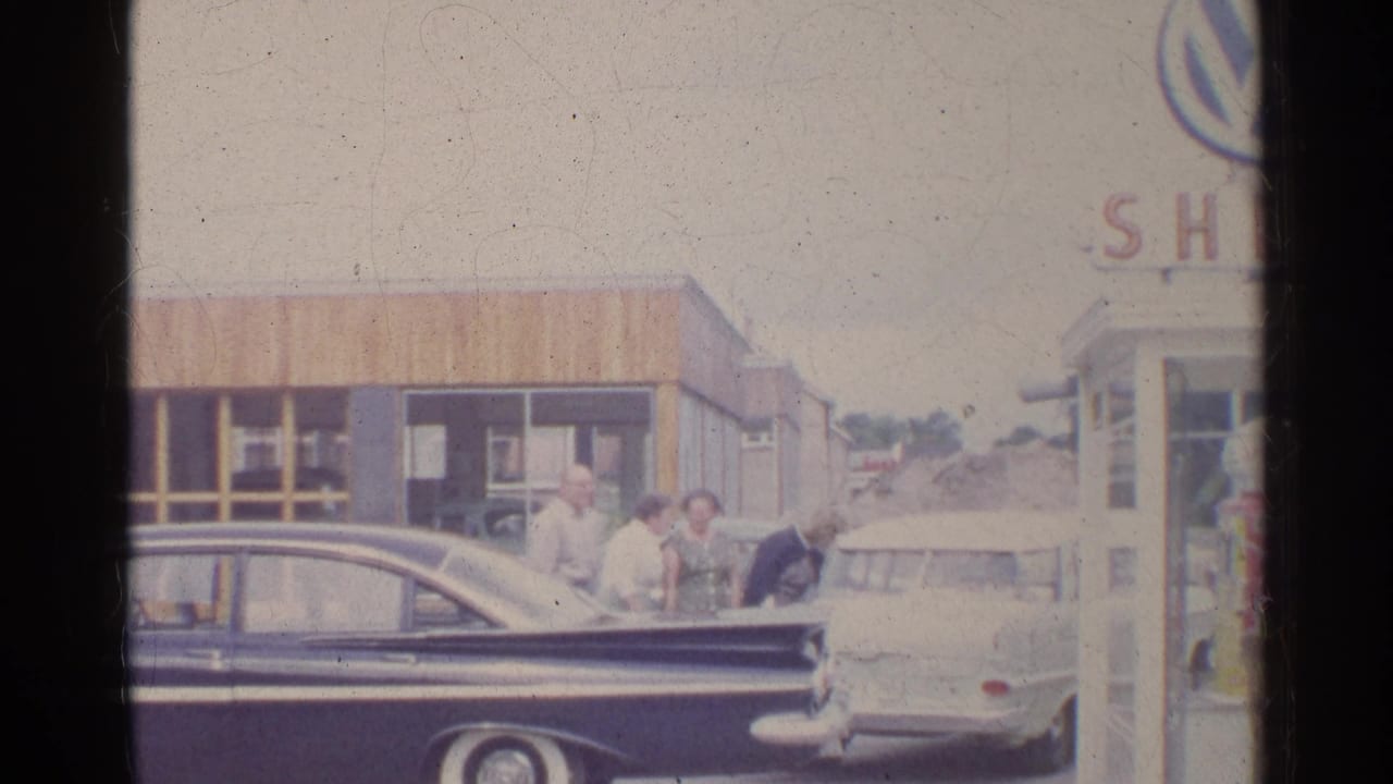 Adults in period clothing stand at a German gas station beside a dark sedan and light car, with partial station branding visible.