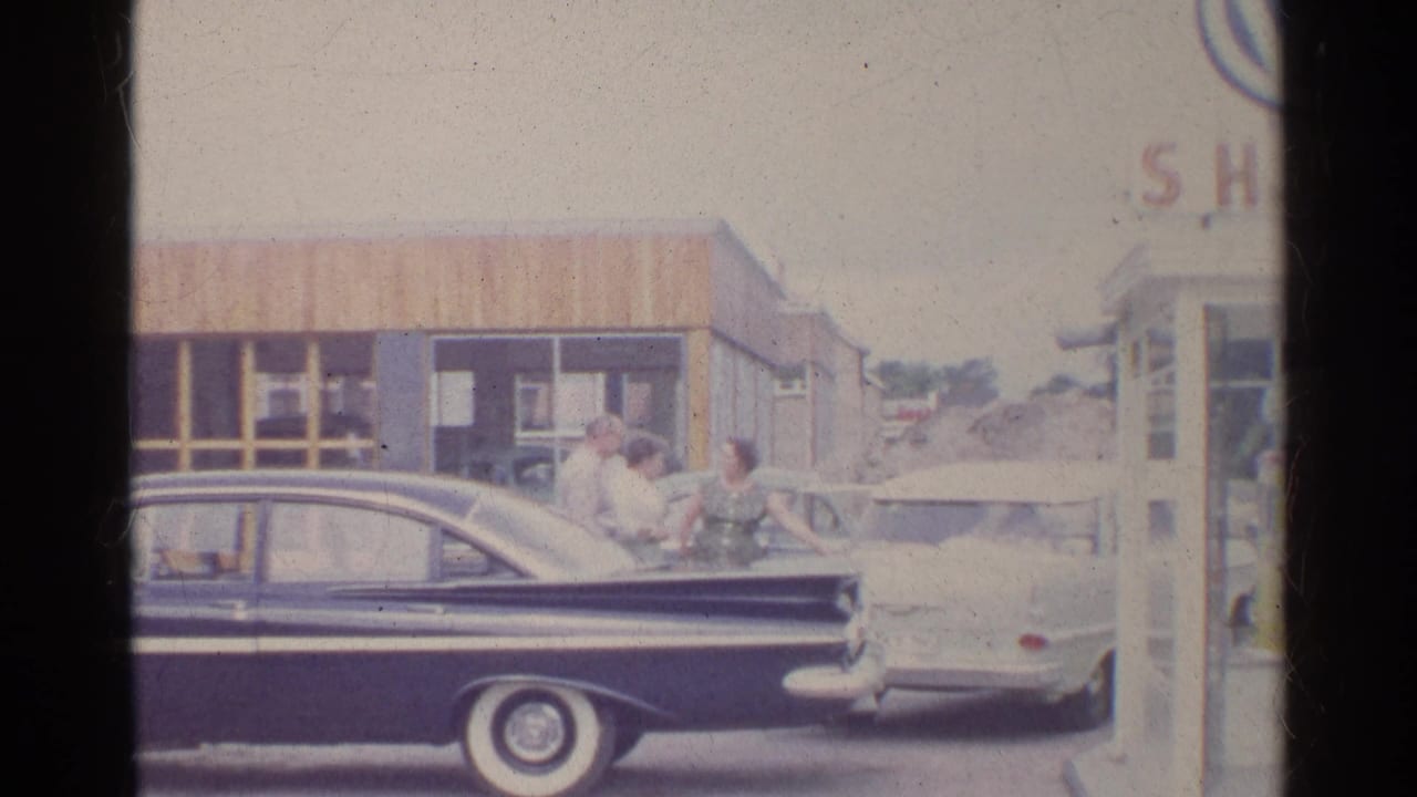 A 1961 Germany roadside scene shows adults beside a dark sedan and light car in front of a small gas station building.