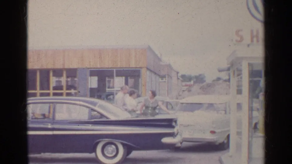 A 1961 Germany roadside scene shows adults beside a dark sedan and light car in front of a small gas station building.