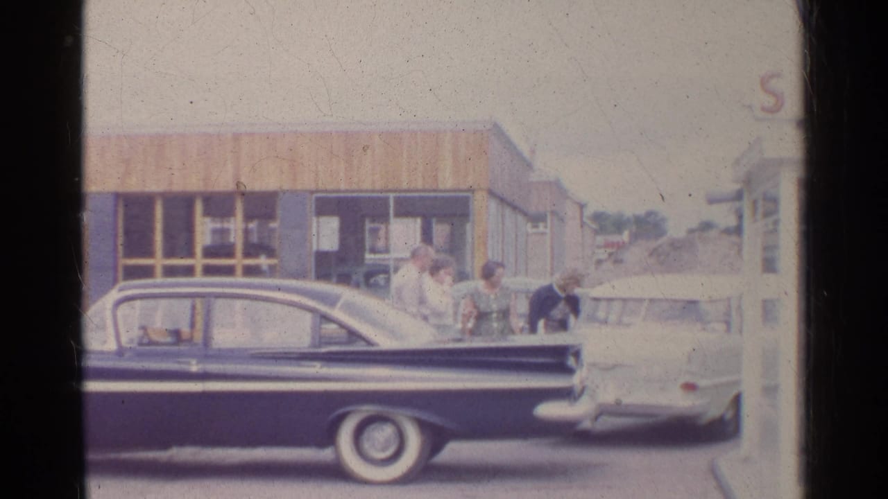 At a German gas station in 1961, adults stand near a dark sedan and light car, with the station building and partial signage behind them.
