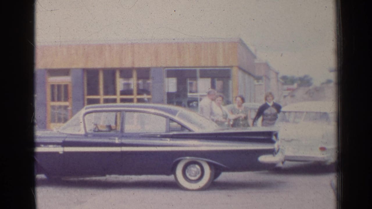Dark blue vintage sedan parked beside adults outside a storefront.