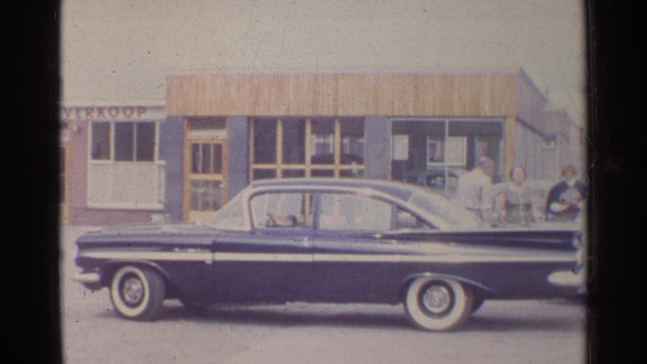Home-movie frame showing the sedan centered outside storefront windows, with a partial VERKOOP sign visible on the building.