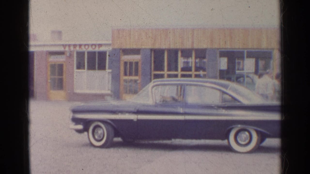 Soft 8mm frame of the dark blue sedan across the lower foreground, with doors, windows and a partial VERKOOP sign behind it.