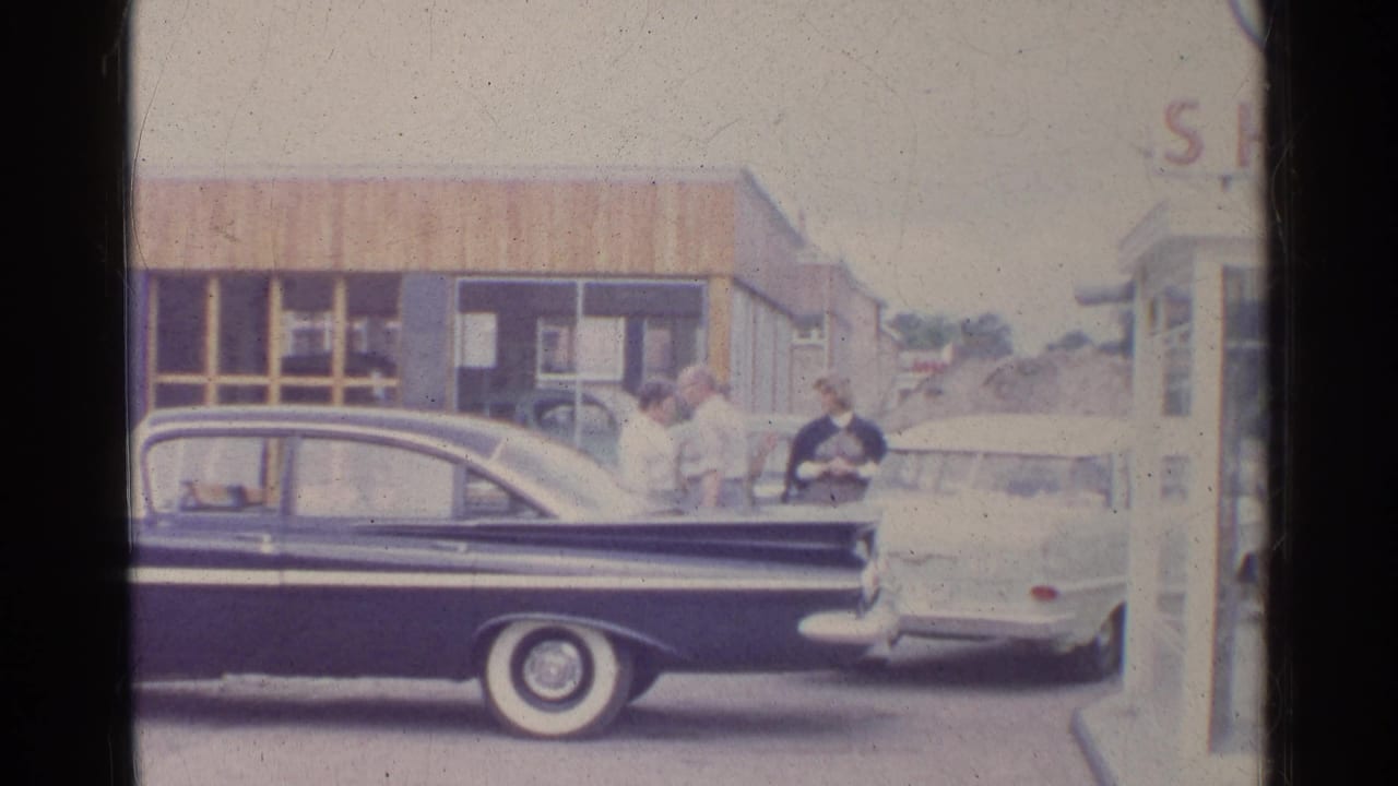 1961 Germany home-movie frame with adults beside two parked cars outside a commercial building, including the dark blue sedan in foreground.