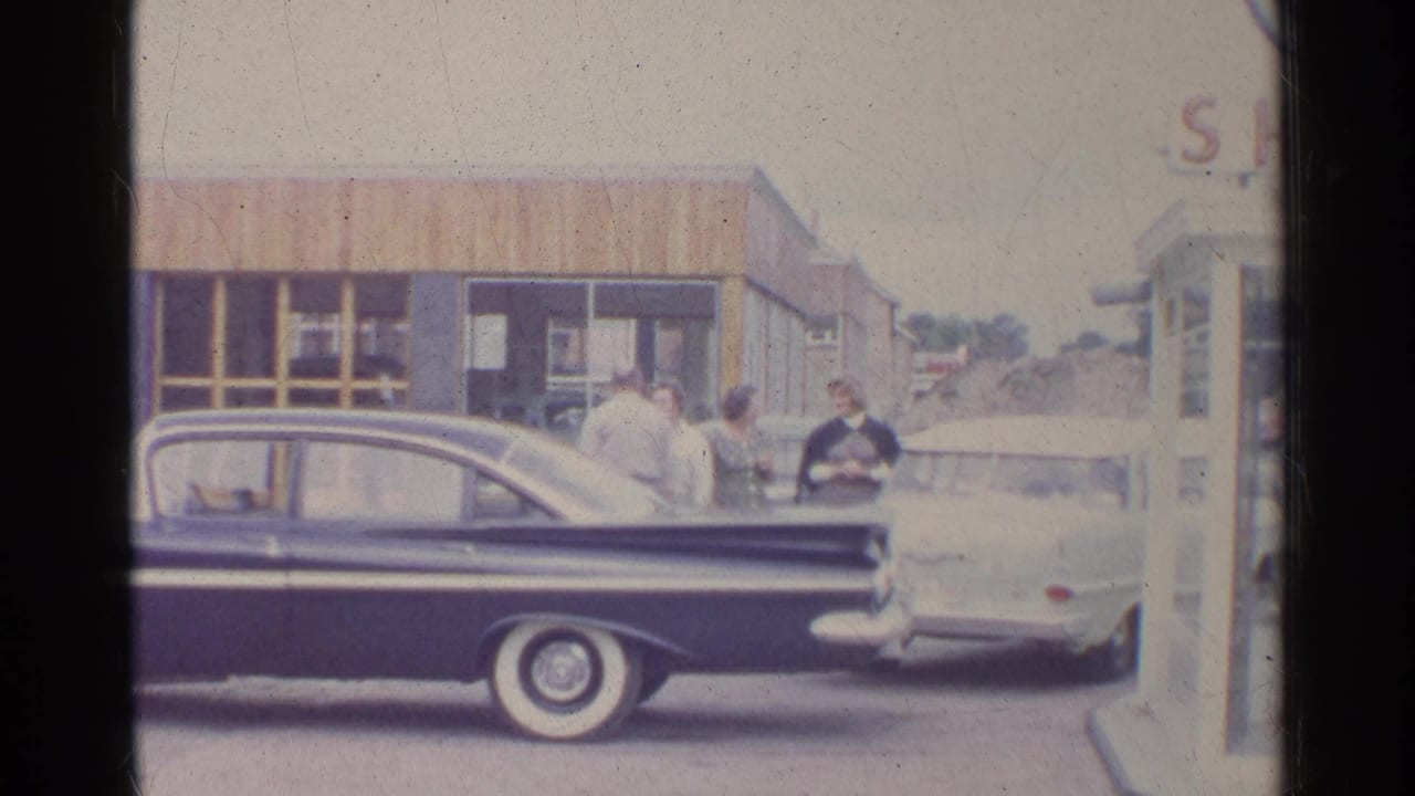 Adults gather near a dark sedan and light car outside a Shell station.