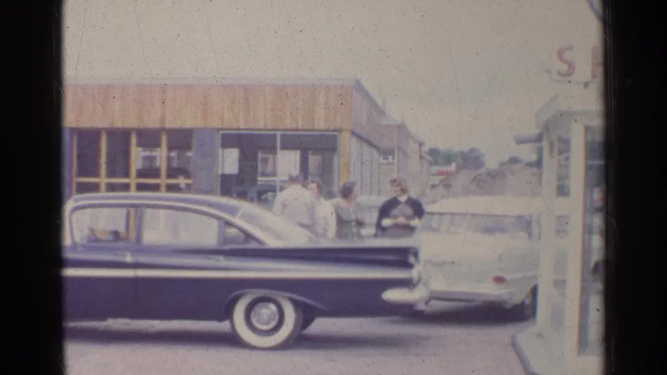 1961 Germany home-movie frame showing adults by a dark sedan and light car at a Shell service station.