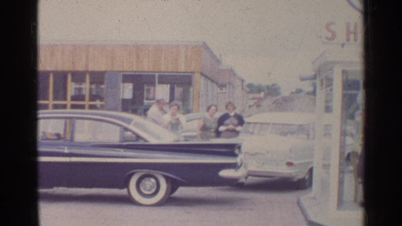 Home-movie frame of a Shell station forecourt with a dark sedan in front and adults near a light car.