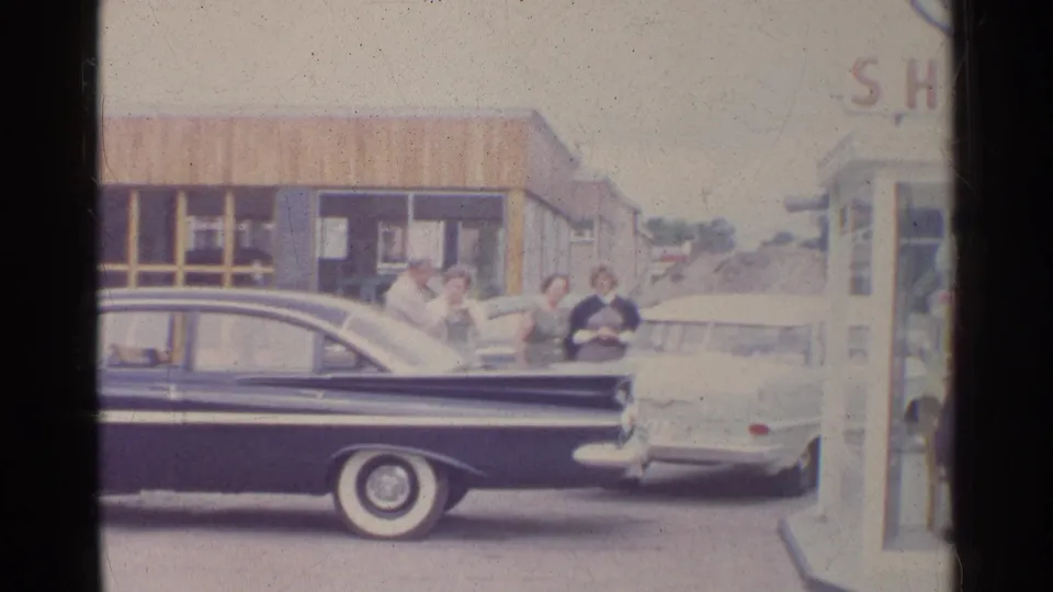 Home-movie frame of a Shell station forecourt with a dark sedan in front and adults near a light car.