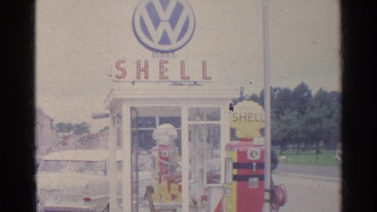 Shell gas station forecourt with a red and yellow pump, white phone booth, Volkswagen sign, and parked white car.