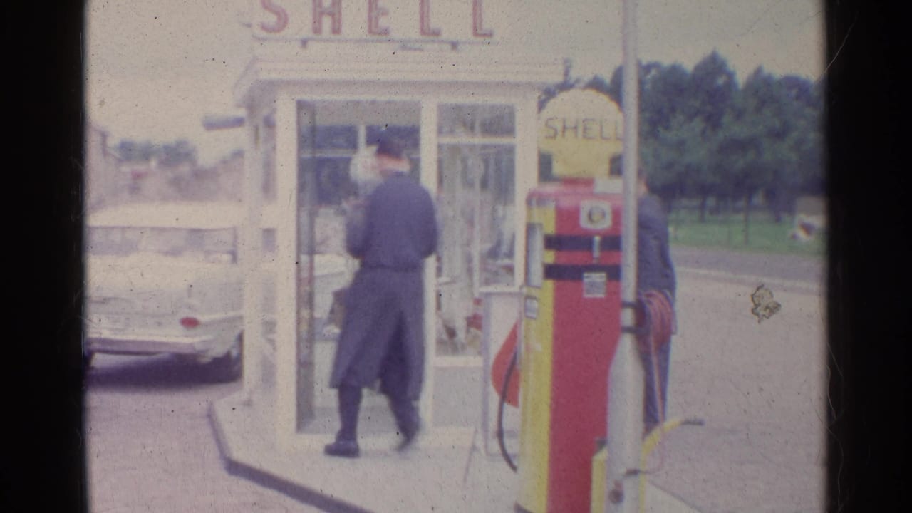 1961 Germany 8mm frame of a man in a dark coat entering or approaching a white phone booth beside a Shell pump at a service station.