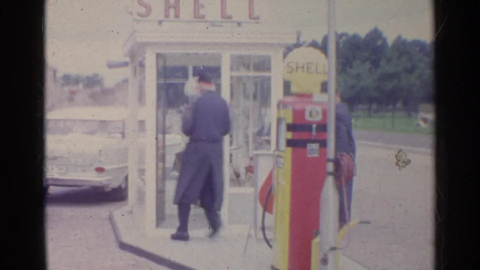 1961 Germany 8mm frame of a man in a dark coat entering or approaching a white phone booth beside a Shell pump at a service station.
