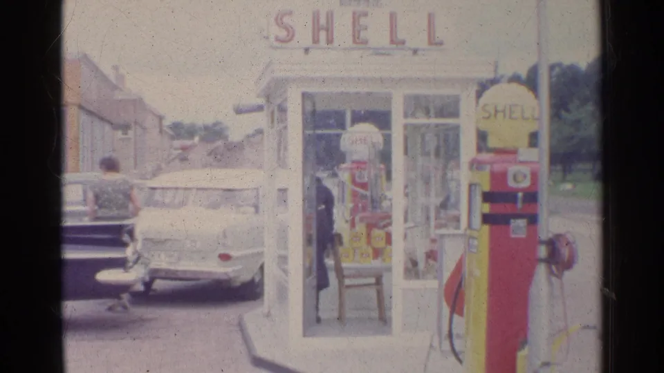 1961 Germany 8mm frame showing the Shell station pump, white phone booth, parked white car, and a person moving near the forecourt.