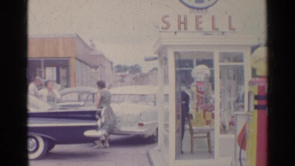 1961 Germany 8mm frame with a woman beside a dark car, a parked white car, and the Shell pump and phone booth in the station forecourt.