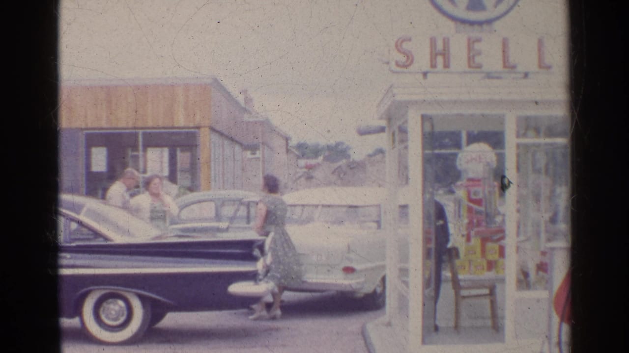 Two women and several men stand near parked sedans outside a Shell station booth.