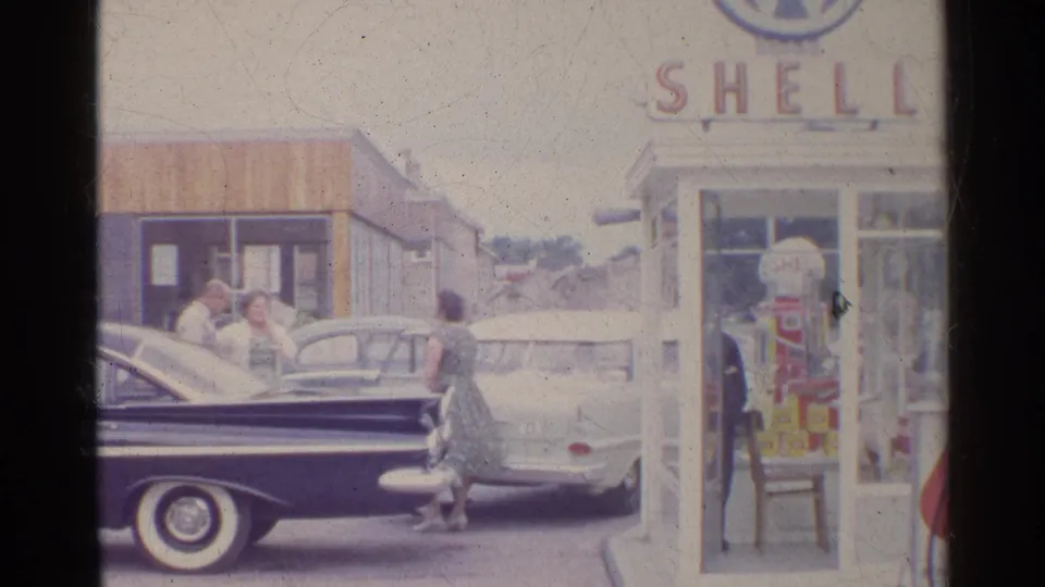 1961 Germany home-movie frame showing adults near parked mid-century cars outside a Shell-branded station booth.