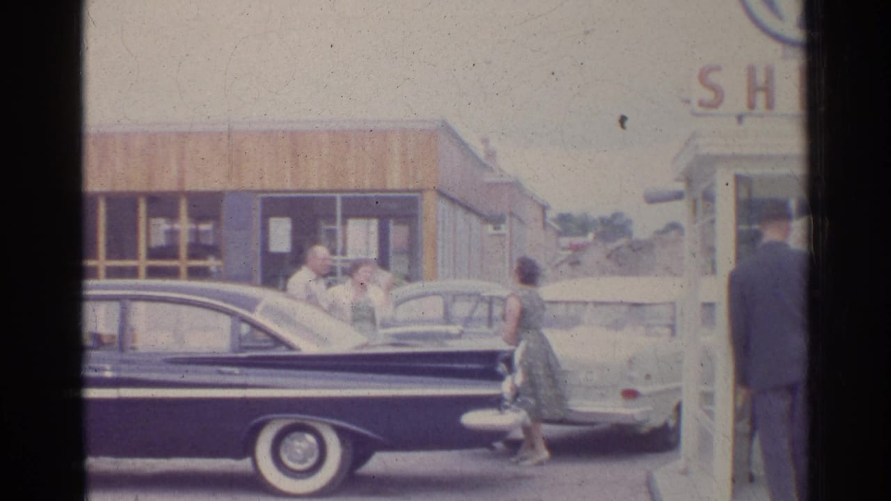 A dark sedan fills the foreground as adults stand near a light car and Shell booth.