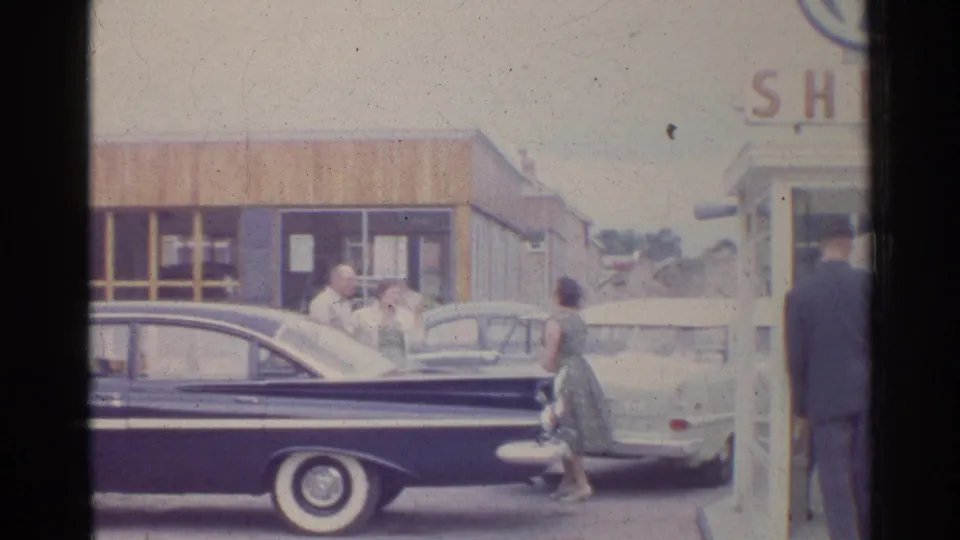 1961 Germany frame with a dark mid-century sedan in front of adults talking near a light car at a Shell station.