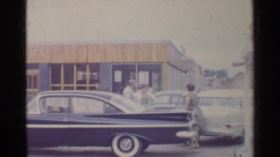 1961 Germany home-movie frame showing adults beside sedans outside a wooden-paneled roadside station building.