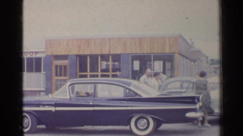 1961 Germany frame showing a dark sedan in the foreground with adults gathered near another car at the station.