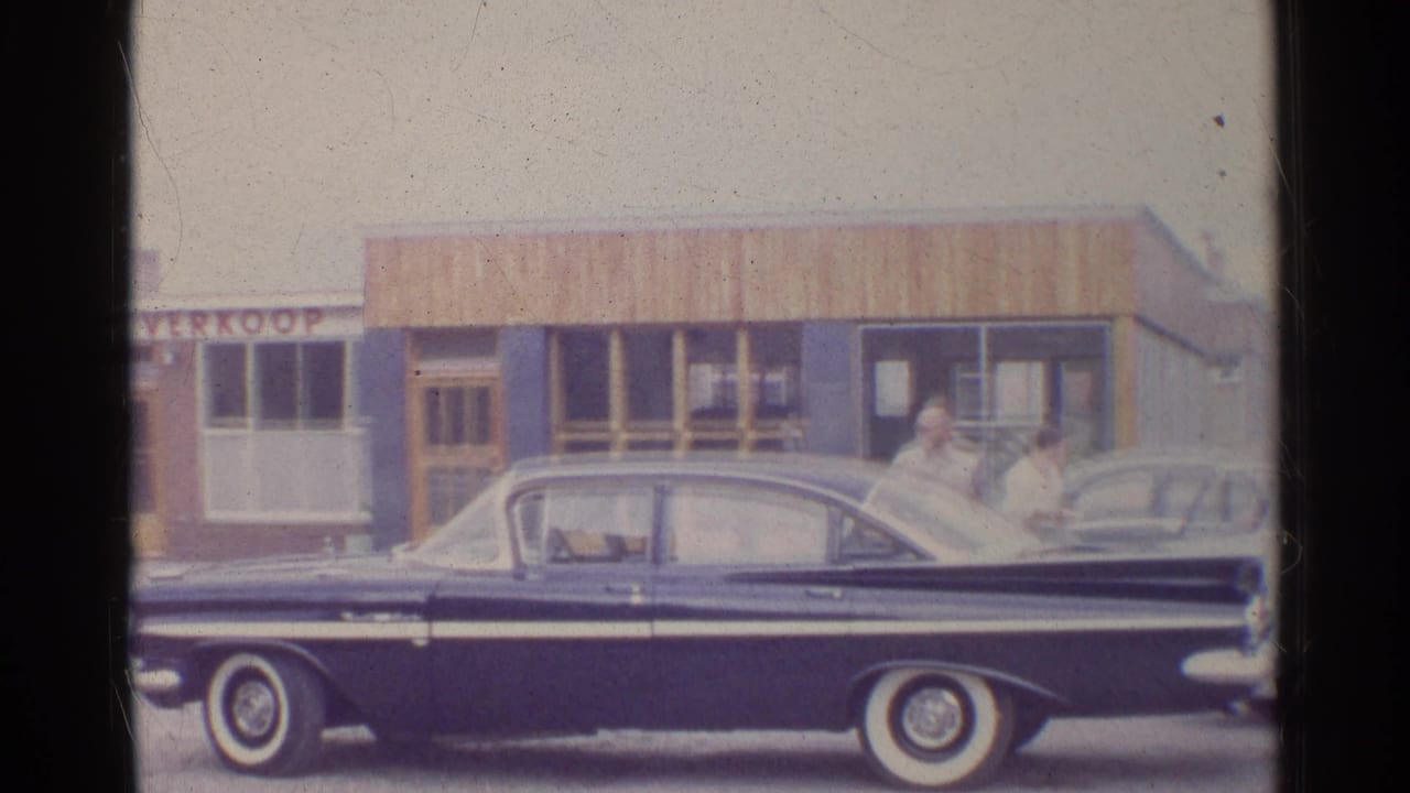 Dark classic sedan parked in front of storefronts with signage and a few adults nearby.