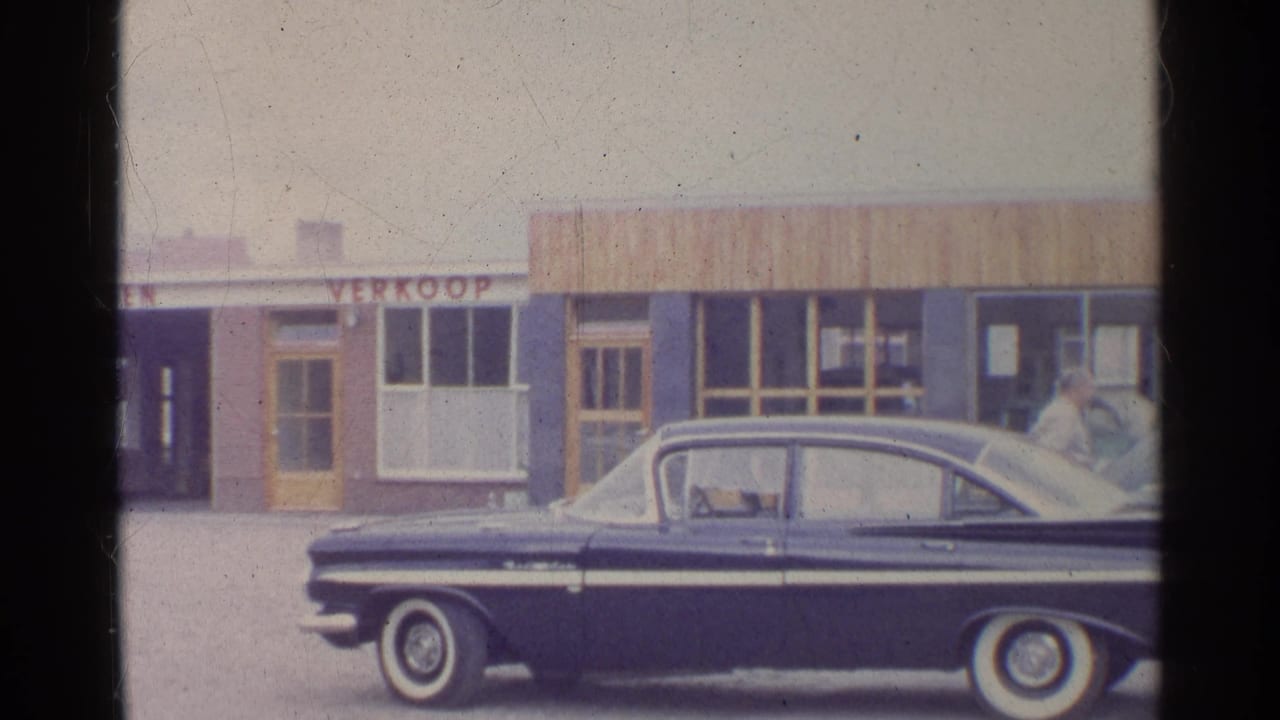 Dark sedan with chrome trim and white-wall tires parked before a German commercial frontage in a 1961 home-movie frame.