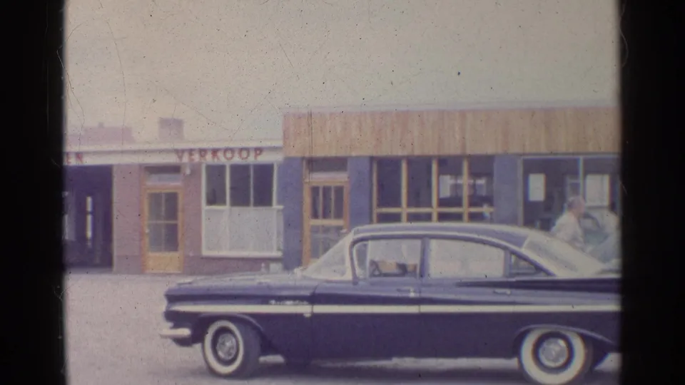 Dark sedan with chrome trim and white-wall tires parked before a German commercial frontage in a 1961 home-movie frame.