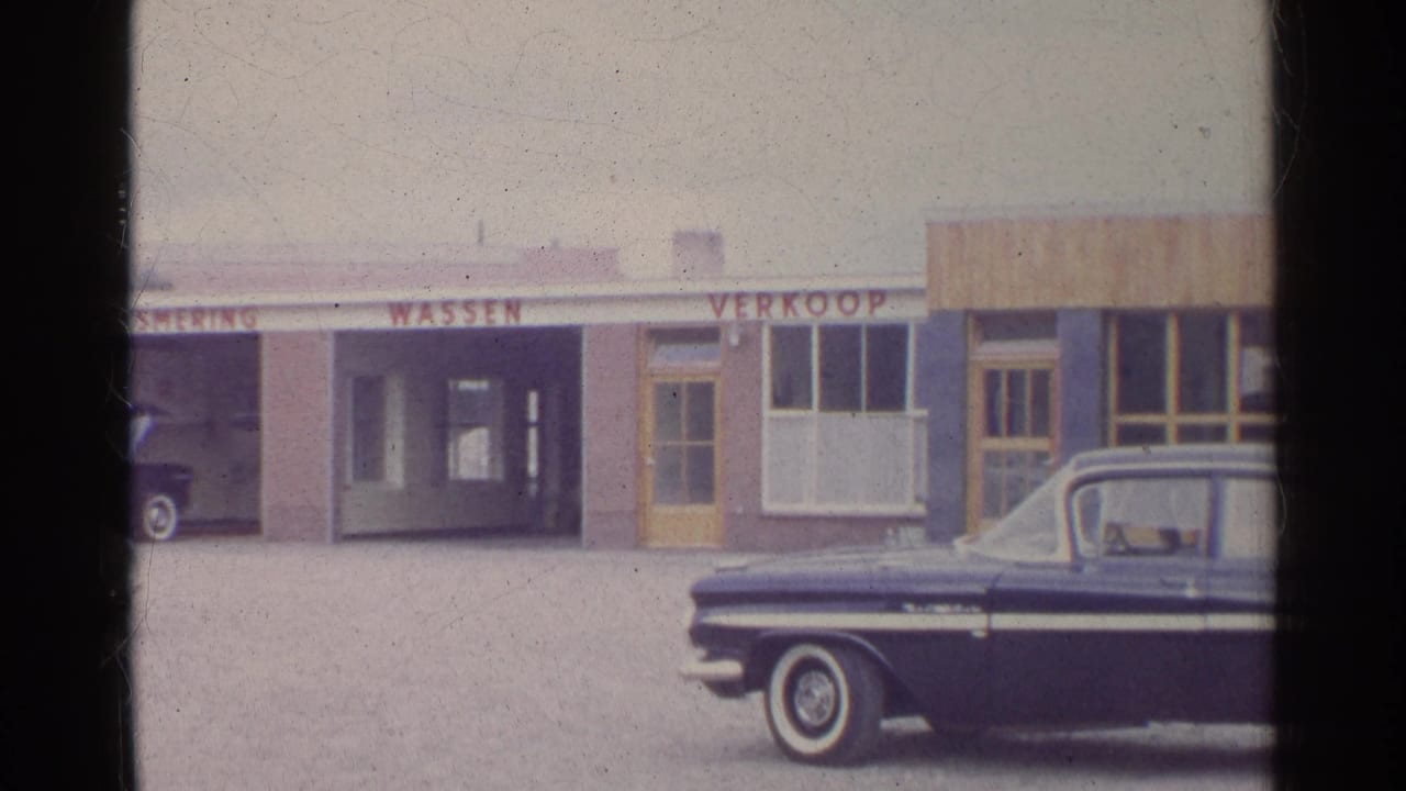 A 1961 Germany-labeled home-movie frame shows the sedan cropped at right before commercial bays marked WASSEN and VERKOOP.