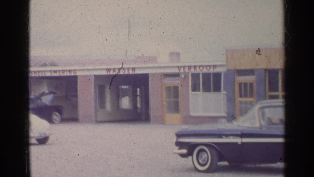 1961 Germany home-movie frame of a Shell service-station frontage with bay signage, parked classic cars, and a dark sedan partly blocking the foreground.
