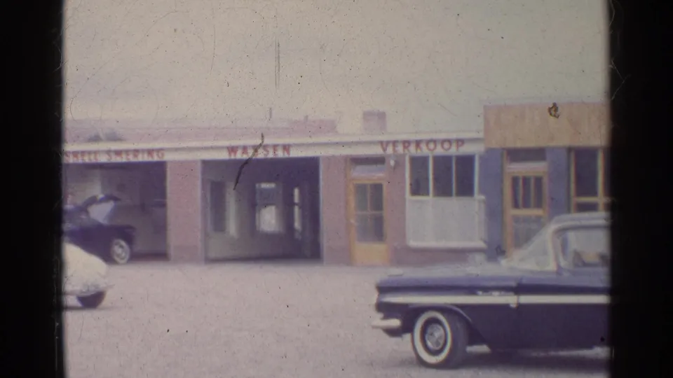 1961 Germany home-movie frame of a Shell service-station frontage with bay signage, parked classic cars, and a dark sedan partly blocking the foreground.