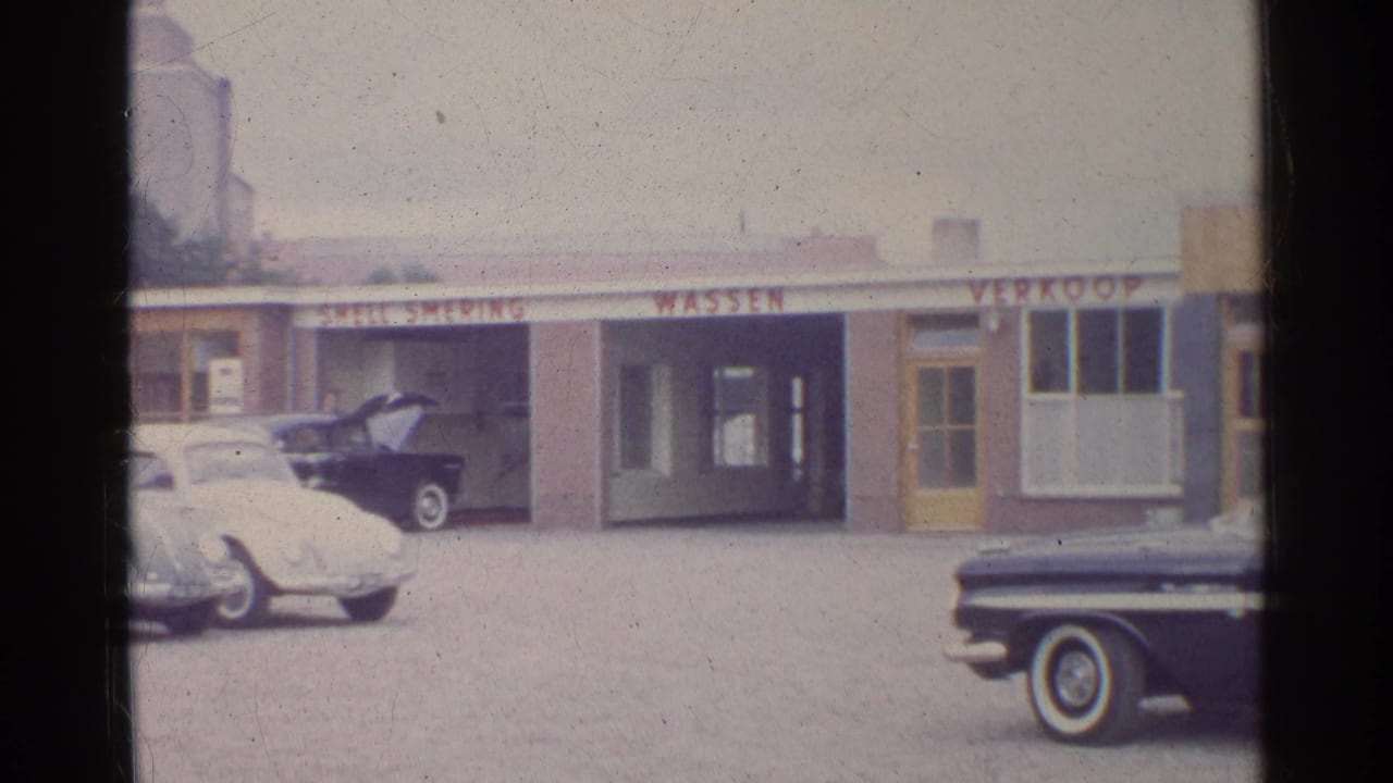 Shell service bays with a white Volkswagen Beetle, a dark car near the garage, and signage along the roofline.