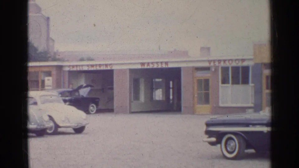 1961 Germany frame showing Shell service bays, wash signage, a white Volkswagen Beetle, and a dark car parked near the garage entrance.