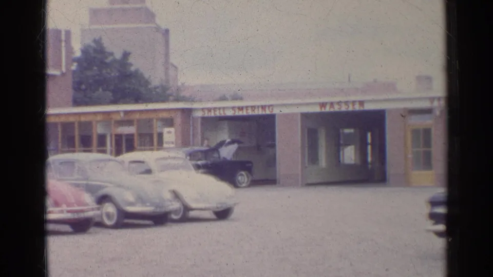 1961 Germany home-movie view of classic cars lined up before a low service-station building with wash signage and storefront windows.