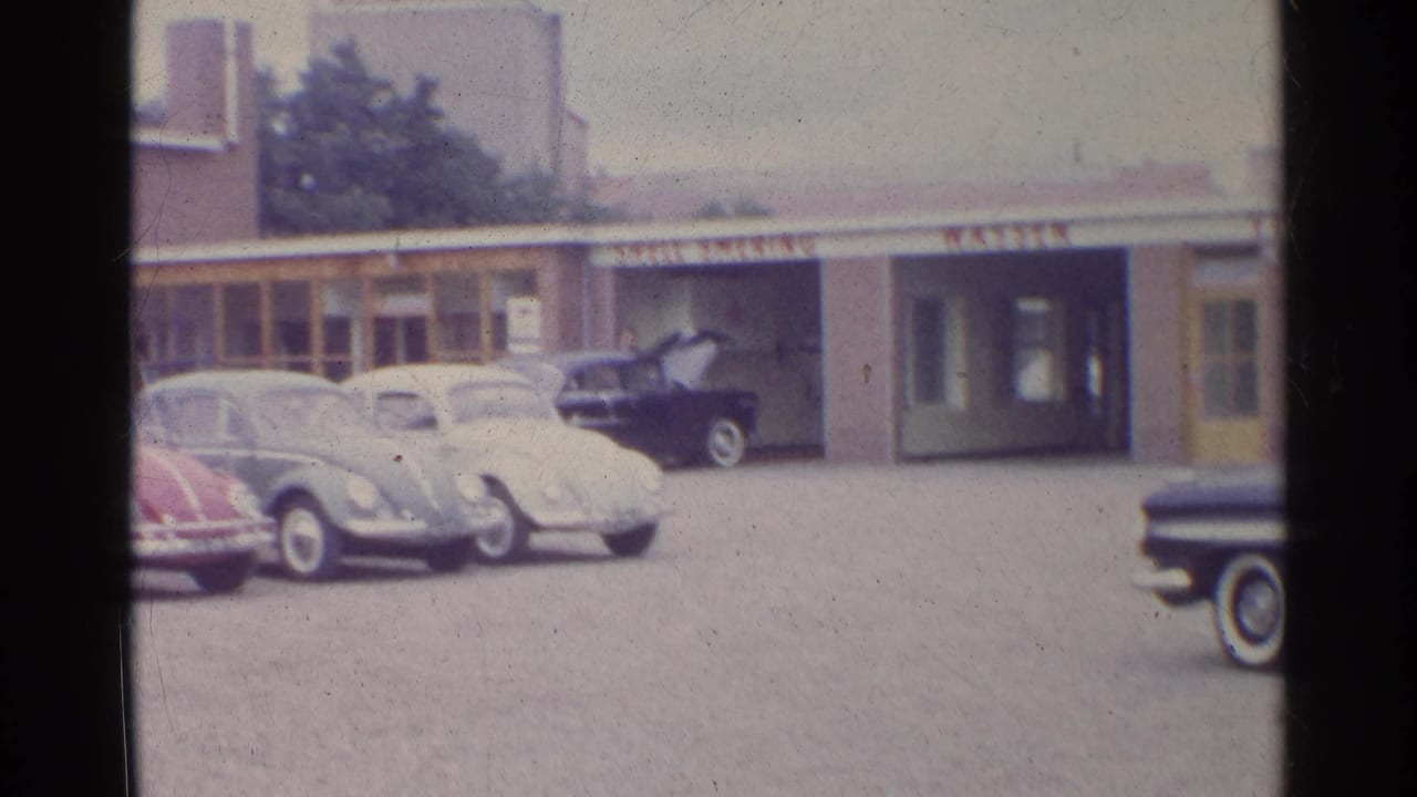 1961 Germany frame with Volkswagen Beetles parked across a Shell station forecourt, a dark car at the service bay, and a sedan at the right edge.