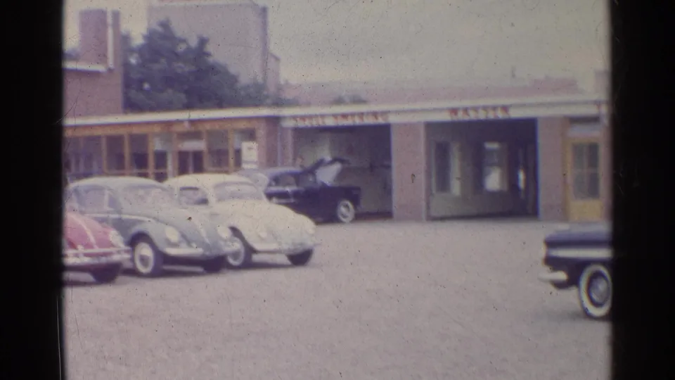 1961 Germany frame with Volkswagen Beetles parked across a Shell station forecourt, a dark car at the service bay, and a sedan at the right edge.