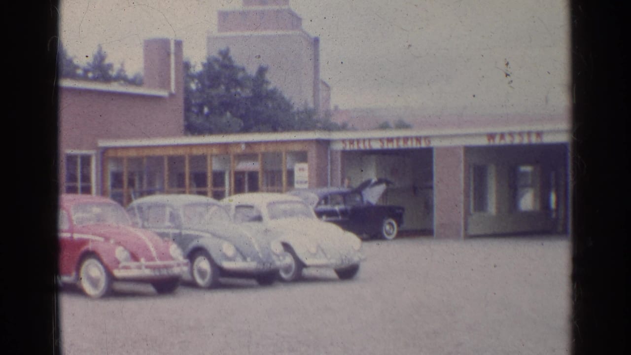 1961 Germany home-movie frame showing red, blue-gray, and white Volkswagen Beetles outside a brown commercial garage building.