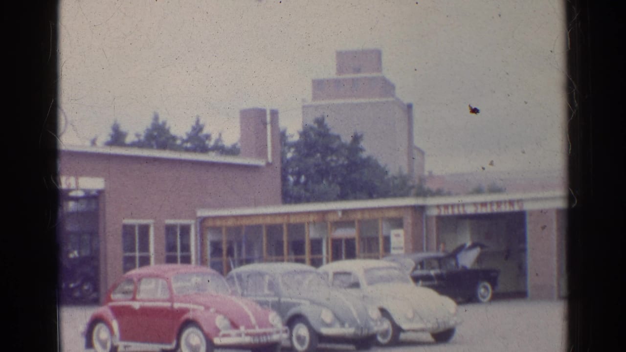 1961 Germany frame with three classic Volkswagen Beetles parked outside a brown roadside garage or commercial building.