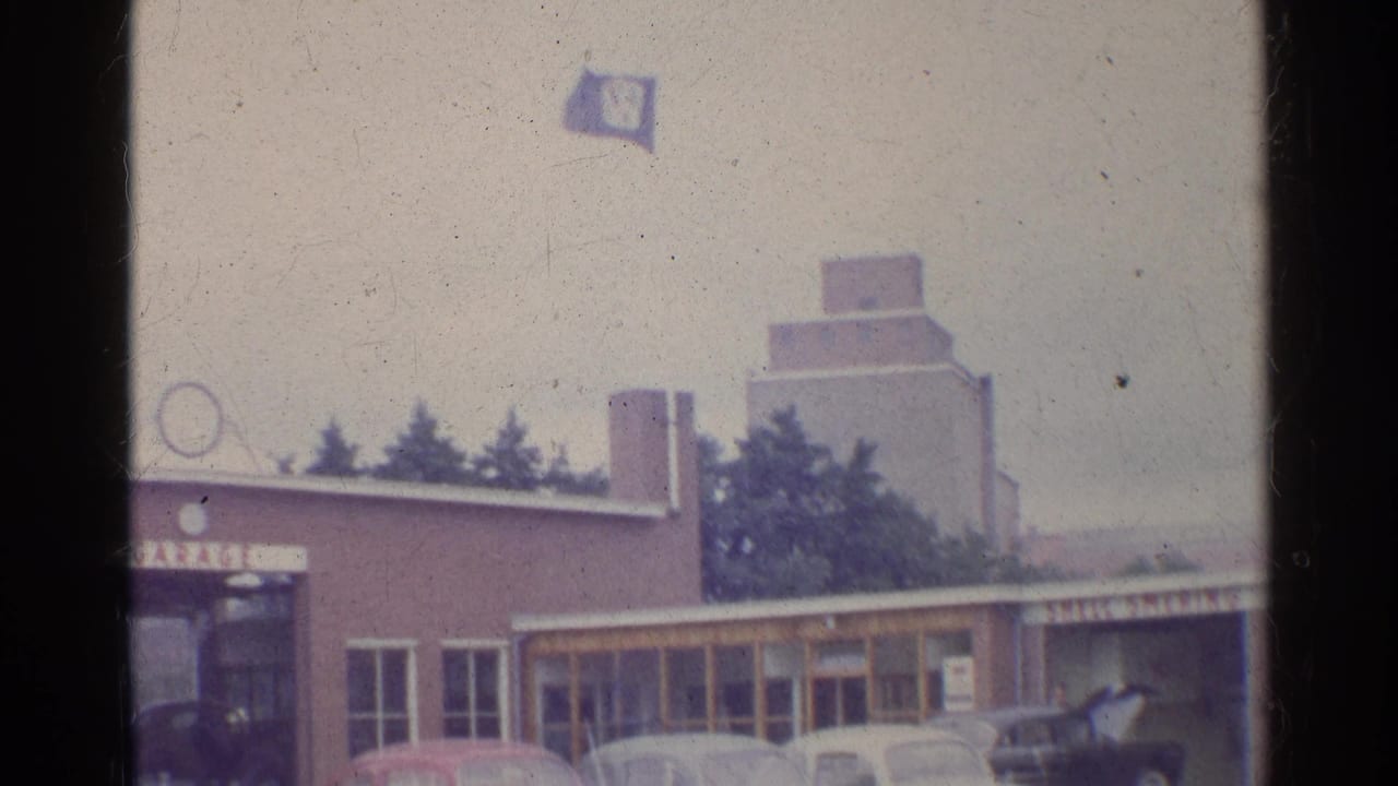 1961 Germany home-movie view of a garage exterior with Beetles, Volkswagen flag, and a larger structure beyond the roofline.