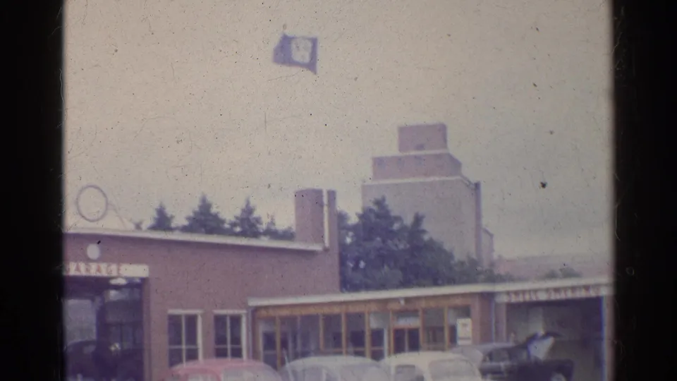 1961 Germany home-movie view of a garage exterior with Beetles, Volkswagen flag, and a larger structure beyond the roofline.