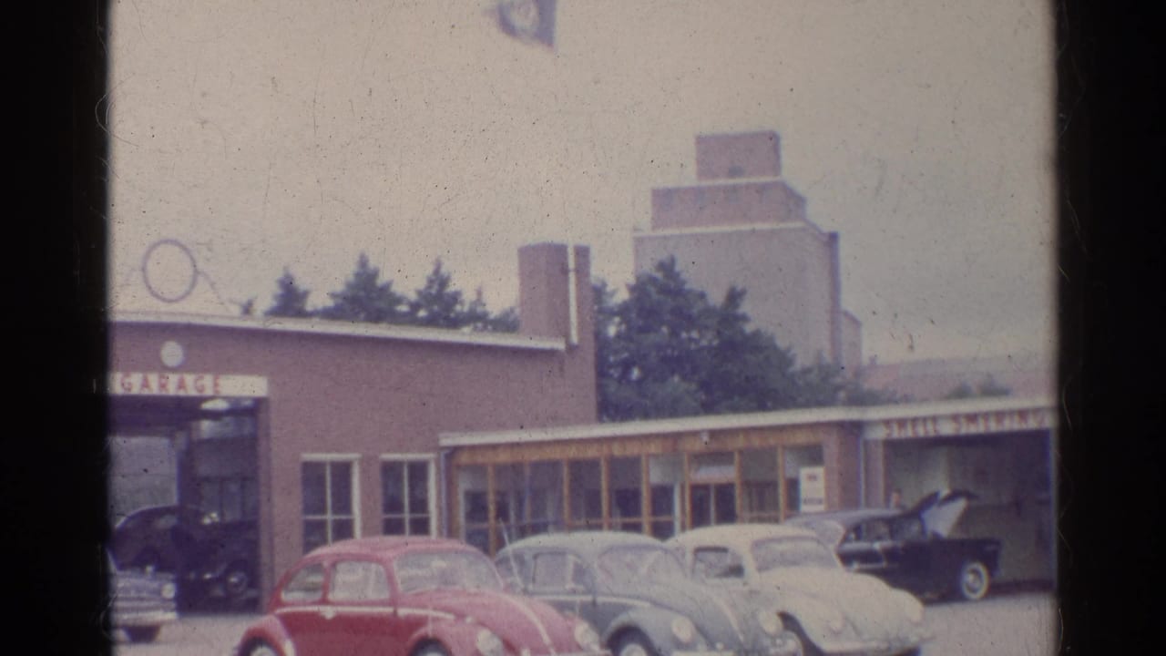 1961 Germany frame showing Beetles parked outside a garage-like building, with a Volkswagen flag and open-hood car visible.