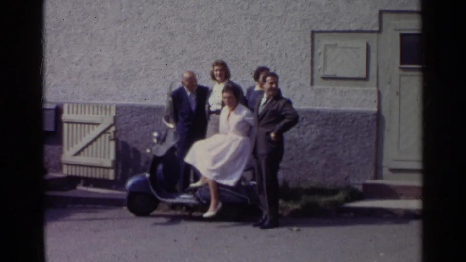 In Germany 1961 home-movie footage, four adults pose around a blue scooter beside a textured exterior wall and pale doorway.