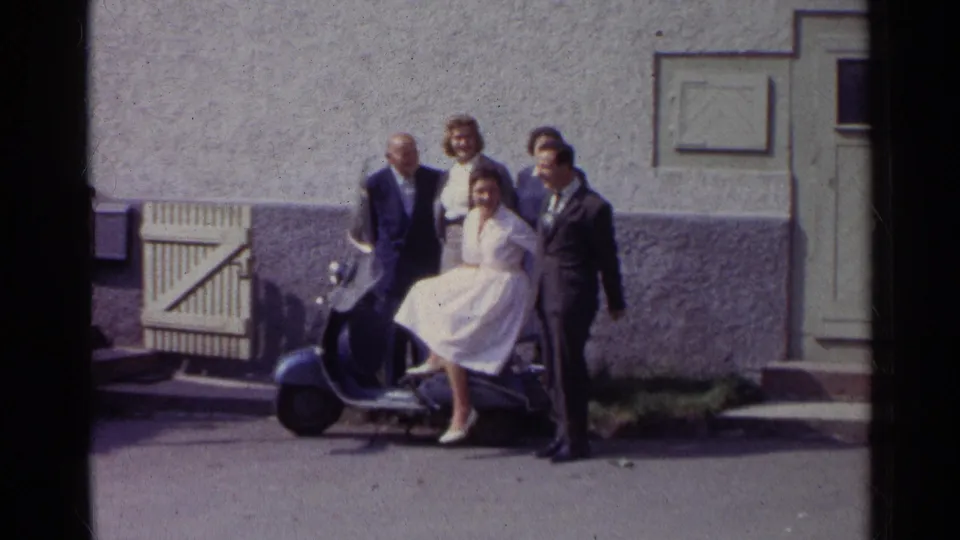 The group remains posed around the scooter, with the seated woman in white centered and adults standing close beside the building.